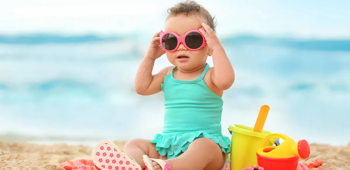 Bebê de maiô verde e óculos rosa sentado na areia, brincando na praia com brinquedos de areia ao lado.