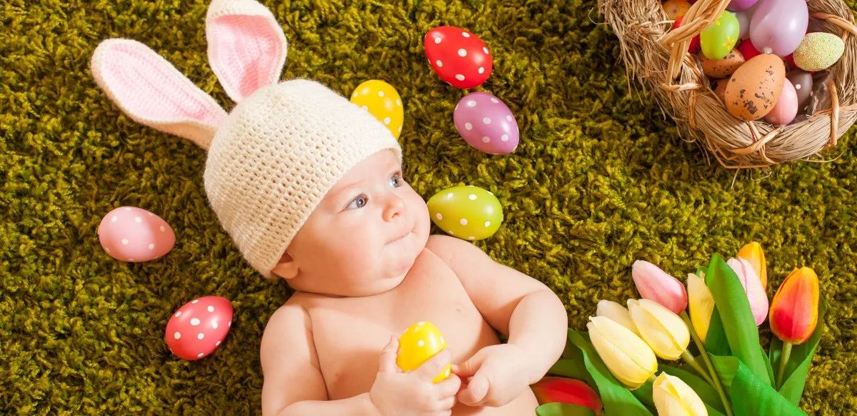 Bebê deitado sobre uma superfície verde usando um gorro de crochê com orelhas de coelho, cercado por ovos de Páscoa coloridos e flores.