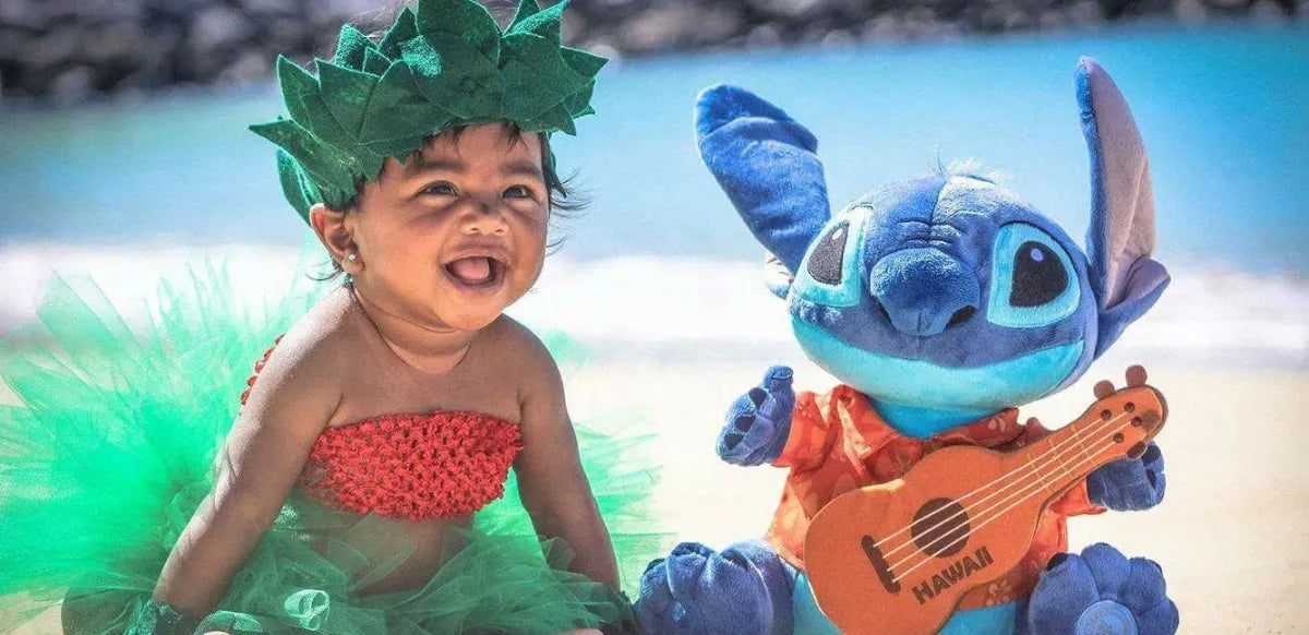 Bebê fantasiada com saia havaiana verde e top vermelho sorrindo ao lado de pelúcia do Stitch com camisa floral e ukulele na praia