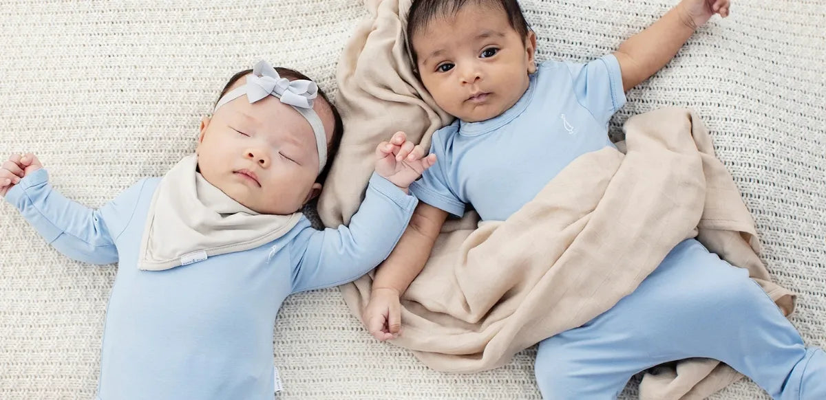 Dois bebês deitados sobre manta clara, vestindo roupinhas azul-claro de fibra de bambu, um dormindo e o outro acordado