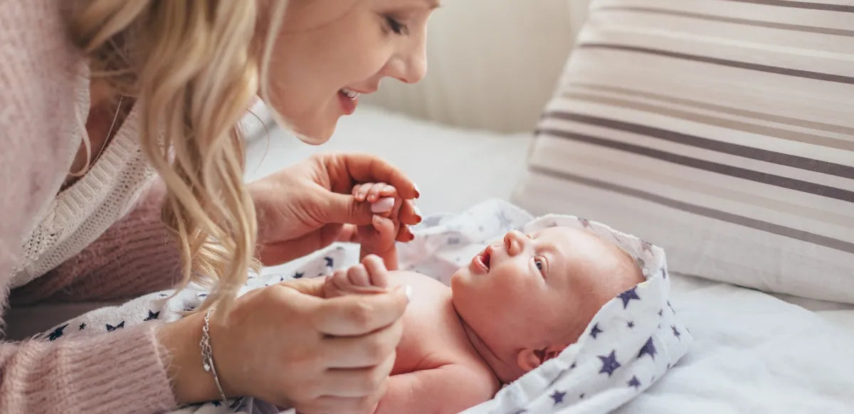 Mãe interagindo com seu bebê recém-nascido, segurando suas mãozinhas e sorrindo carinhosamente enquanto ele a observa deitado em uma cama aconchegante.