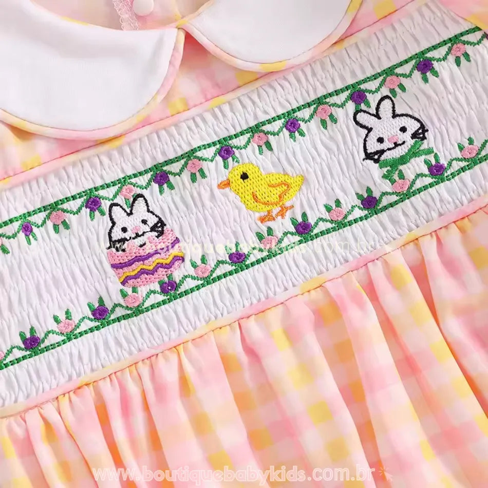 Bordado temático de Páscoa no peito do body, com coelhinhos, pintinho e detalhes florais em ponto smock. Tons vibrantes e acabamento sofisticado.