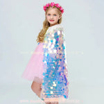 Menina loira usando a Capa Infantil Temática Sereia Encantada, com tecido holográfico brilhante e detalhes de gola rendada branca e fita dourada.