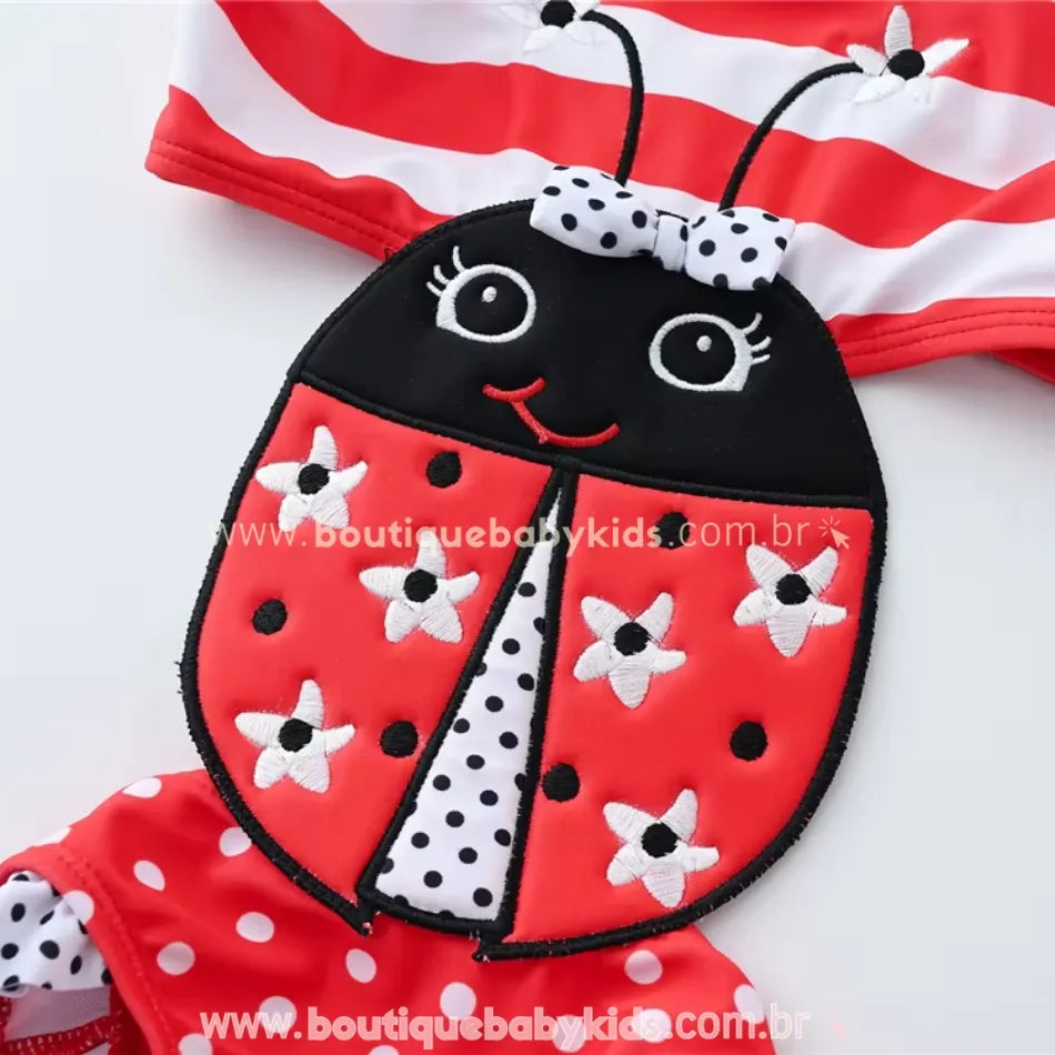 Close da estampa de joaninha do maiô infantil engana mamãe, com anteninhas decorativas, laço de bolinhas e aplicações bordadas de flores brancas.