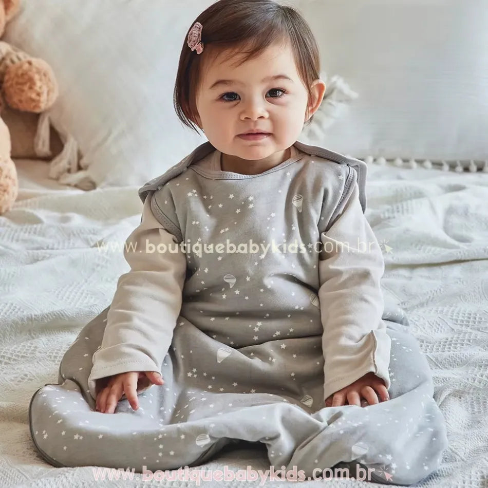 Bebê sentado na cama vestindo o saco de dormir cinza com estampa de estrelas e balões, com mangas longas por baixo, em um ambiente aconchegante com pelúcia ao fundo.
