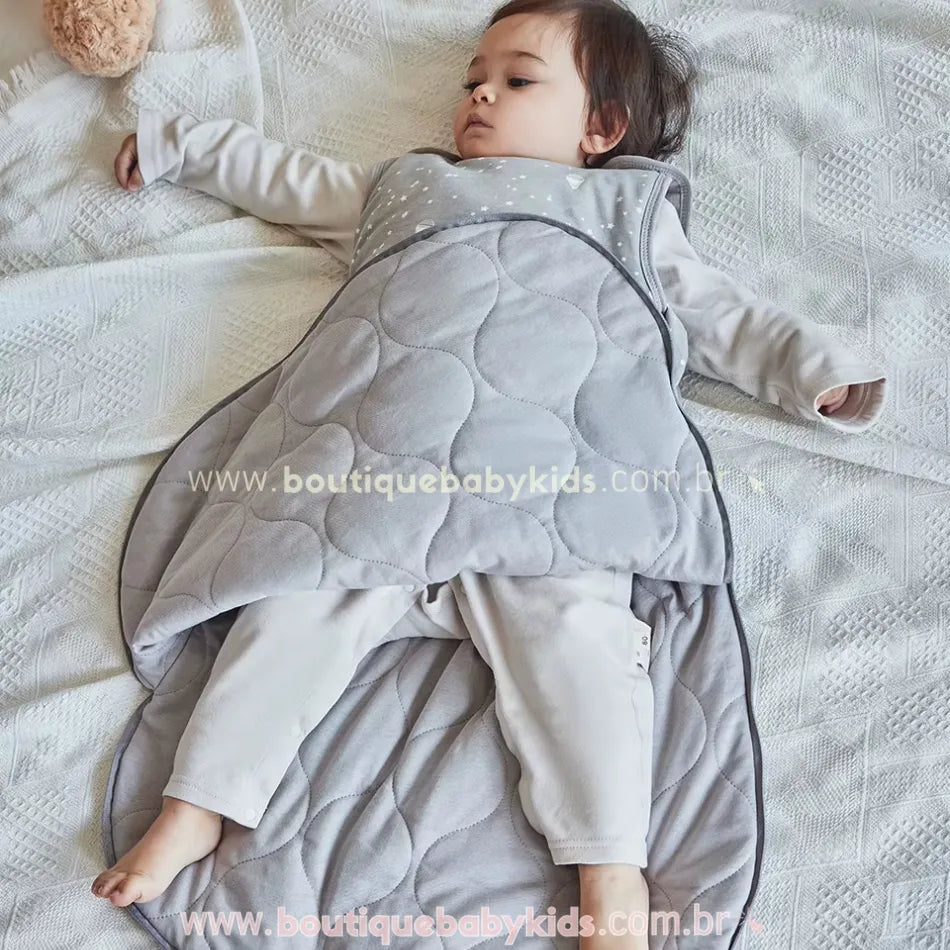 Bebê deitado sobre a cama parcialmente coberto pelo saco de dormir, mostrando o forro acolchoado e o fechamento lateral por zíper.