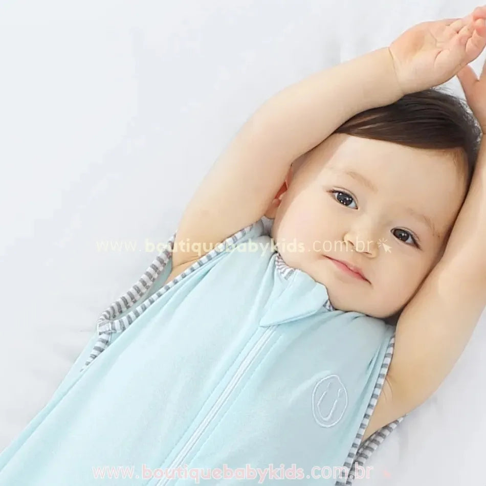 Bebê deitado e sorrindo, com os braços levantados acima da cabeça, vestindo o saco de dormir azul claro com bordado discreto de smile.