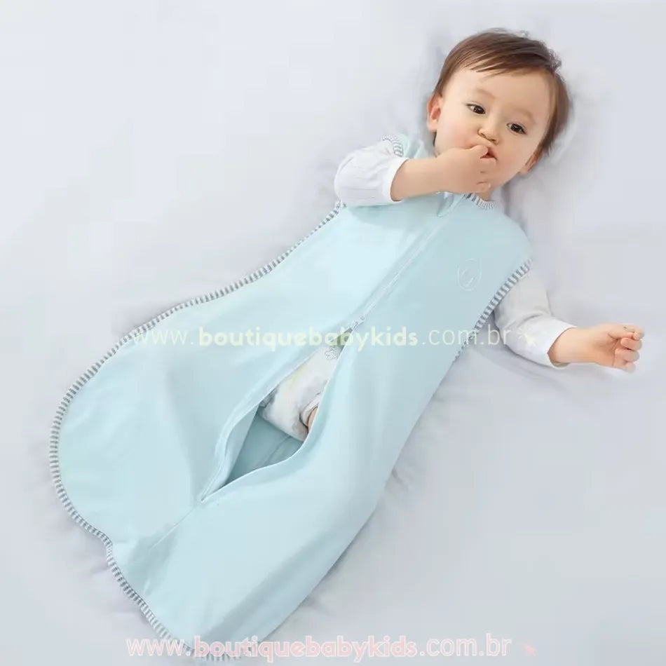 Bebê deitado em um saco de dormir azul claro, com zíper central e bordas com acabamento listrado. O bebê segura a parte inferior do saco, mostrando sua flexibilidade.