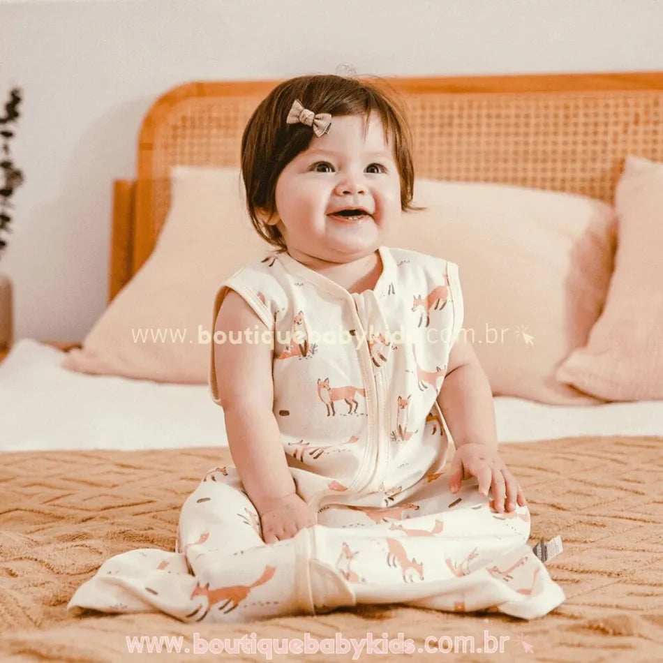 Bebê sorrindo e sentado na cama, usando o saco de dormir com estampa temática de raposas e fundo neutro, proporcionando conforto e liberdade de movimentos.