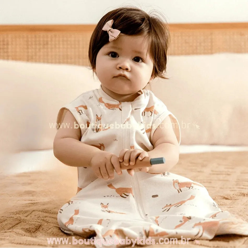 Bebê sentado na cama, segurando um brinquedo e vestindo o saco de dormir estampado com raposas, com abertura frontal em zíper.