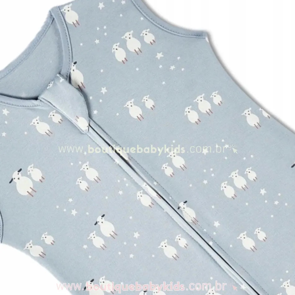 Close-up do tecido azul claro do saco de dormir para bebê, com estampa delicada de carneirinhos brancos e pequenas estrelas. O zíper frontal com proteção no topo também é visível.