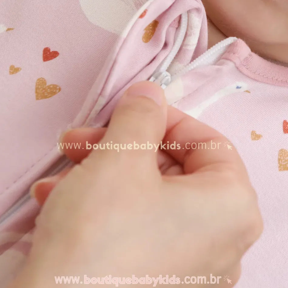 Close-up do zíper do saco de dormir para bebê, com aba protetora para evitar contato com a pele. Modelo rosa com estampa de cisnes e pequenos corações.