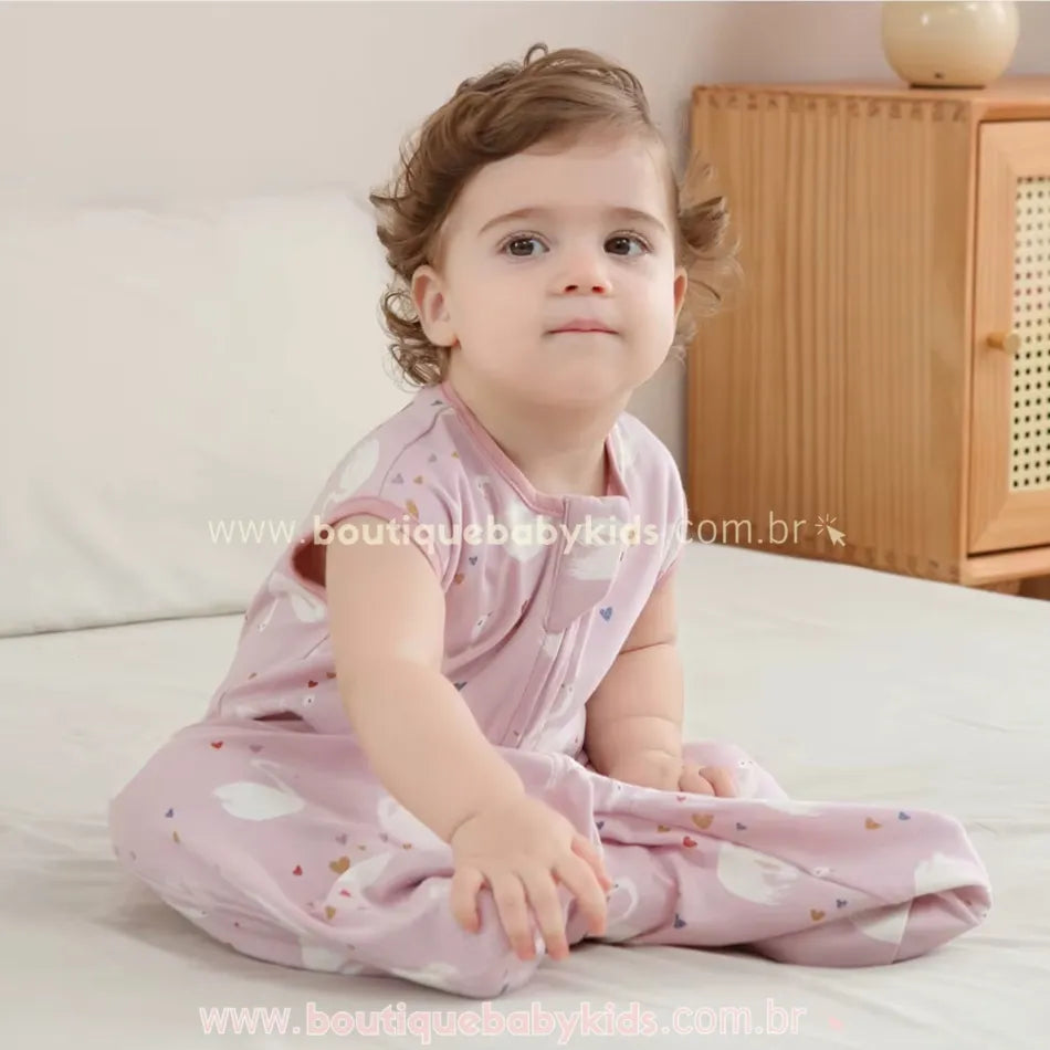 Bebê usando o saco de dormir rosa com estampa de cisnes, mostrando o caimento confortável da peça. Modelo sem mangas, feito 100% em algodão.