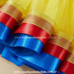 Close-up da barra da saia infantil Branca de Neve com camadas de fita de cetim azul e vermelho, acabamento dourado brilhante para um toque encantador.