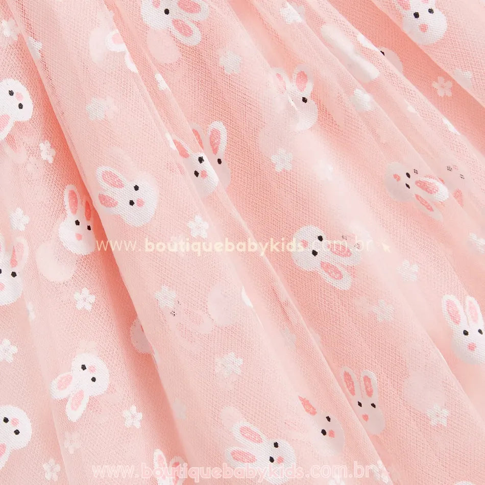 Close na saia do vestido rosa, evidenciando o tule leve e fluído com estampa de coelhinhos brancos e detalhes de pequenas flores, ideal para um visual delicado e festivo. 