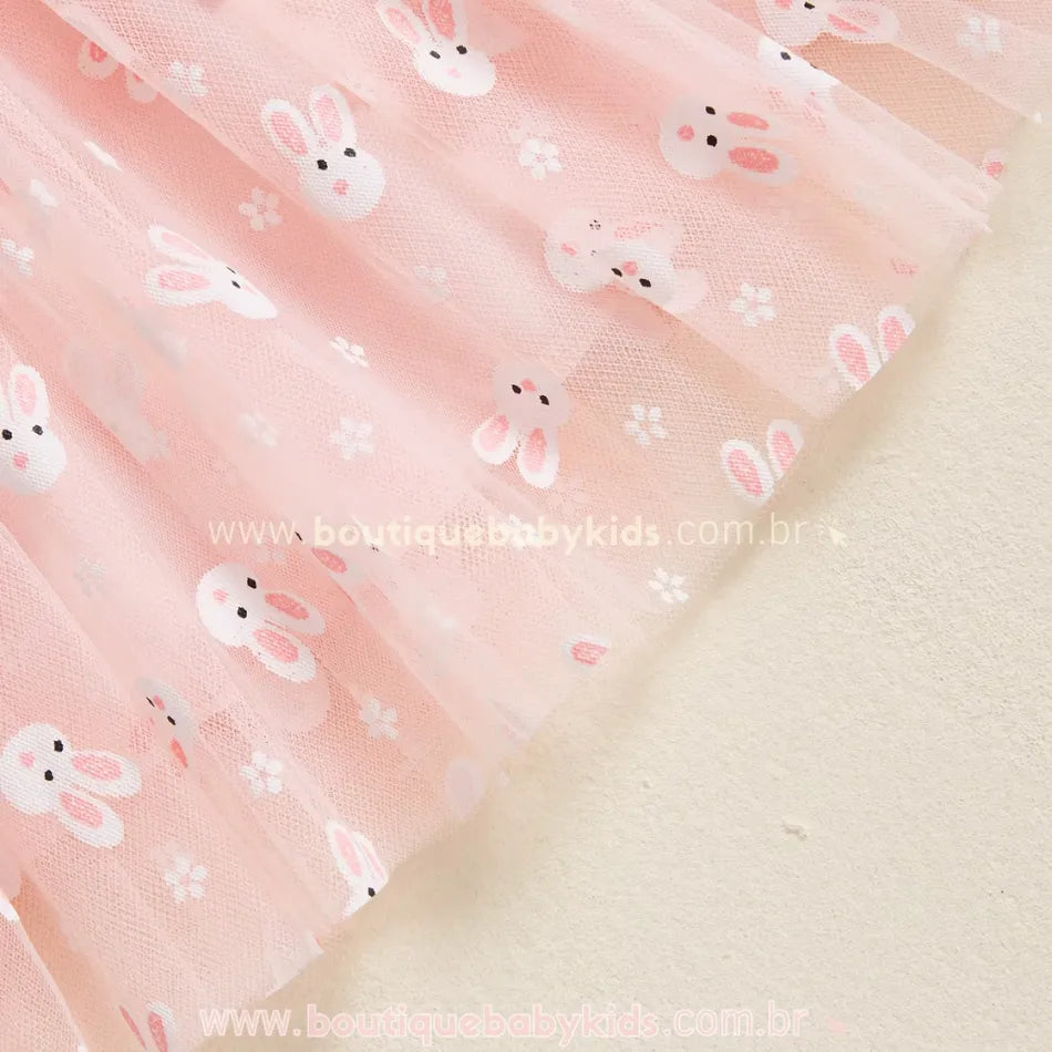 Vista detalhada da barra do vestido rosa, mostrando a transparência do tule e a suavidade da estampa de coelhinhos, conferindo um acabamento delicado à peça. 