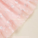 Vista detalhada da barra do vestido rosa, mostrando a transparência do tule e a suavidade da estampa de coelhinhos, conferindo um acabamento delicado à peça. 