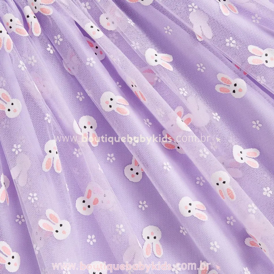 Close na saia do vestido lilás, mostrando a leveza do tecido e os detalhes da estampa de coelhinhos brancos e flores, conferindo um toque lúdico à peça. 