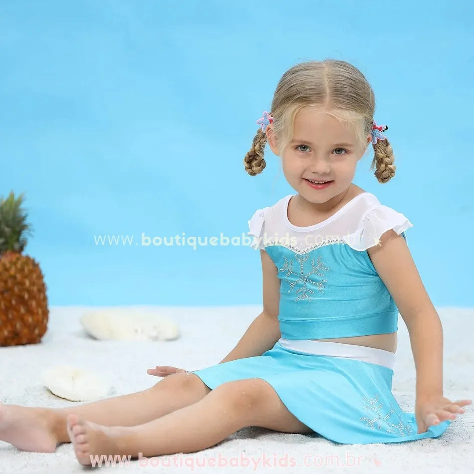 Menina sentada na areia usando o biquíni inspirado na Elsa, com expressão alegre e detalhes brilhantes do conjunto.