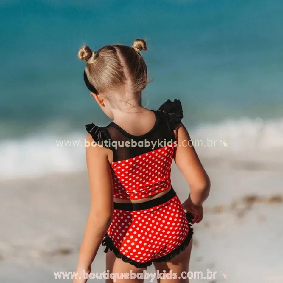 Menina de costas na praia vestindo o biquíni infantil poá minnie mouse, com detalhe da transparência preta no top e babados no short.