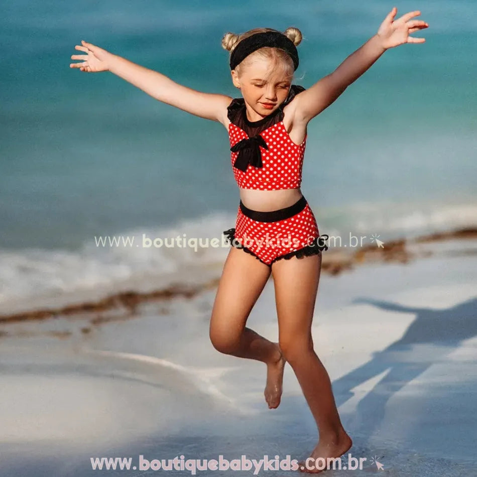 Criança correndo na areia usando o biquíni infantil poá minnie mouse vermelho, com babados nas laterais do short.