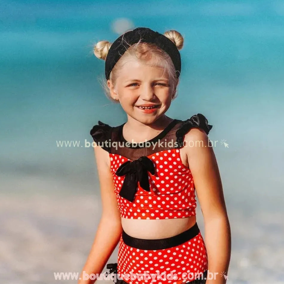 Menina sorridente na praia usando o biquíni infantil poá minnie mouse vermelho com laço preto e mangas franzidas.
