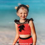 Menina sorridente na praia usando o biquíni infantil poá minnie mouse vermelho com laço preto e mangas franzidas.