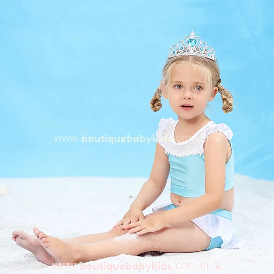 Criança sentada na areia vestindo o biquíni infantil Princesa Cinderela, com top de mangas curtas e babados e shortinho com saia embutida, enquanto brinca.