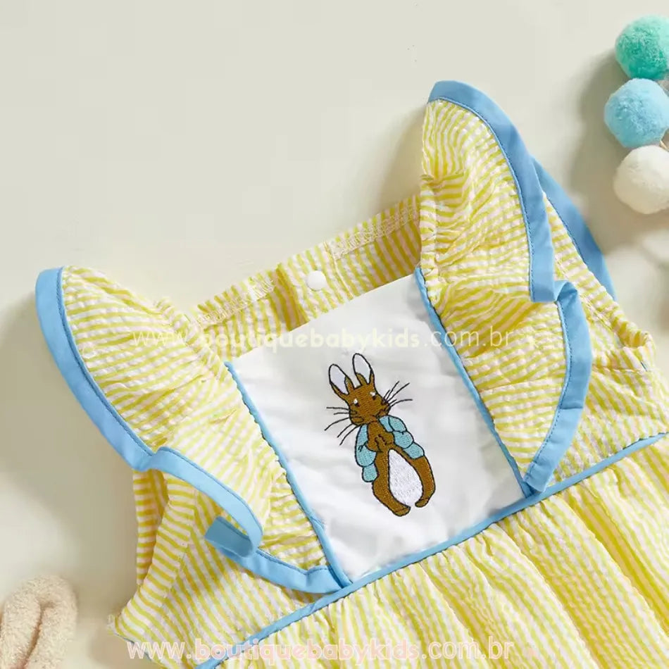 Detalhe do bordado frontal do body de bebê, com coelhinho delicadamente costurado sobre tecido branco, rodeado por babados amarelos e azuis.