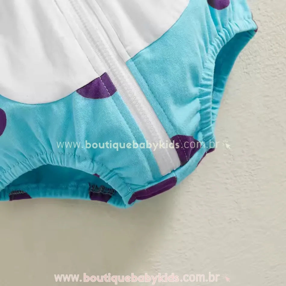 Detalhe da parte inferior do body bebê fantasia Monstrinho Sulley, destacando o tecido azul estampado com manchas roxas, zíper frontal branco e acabamento elástico nas perninhas.