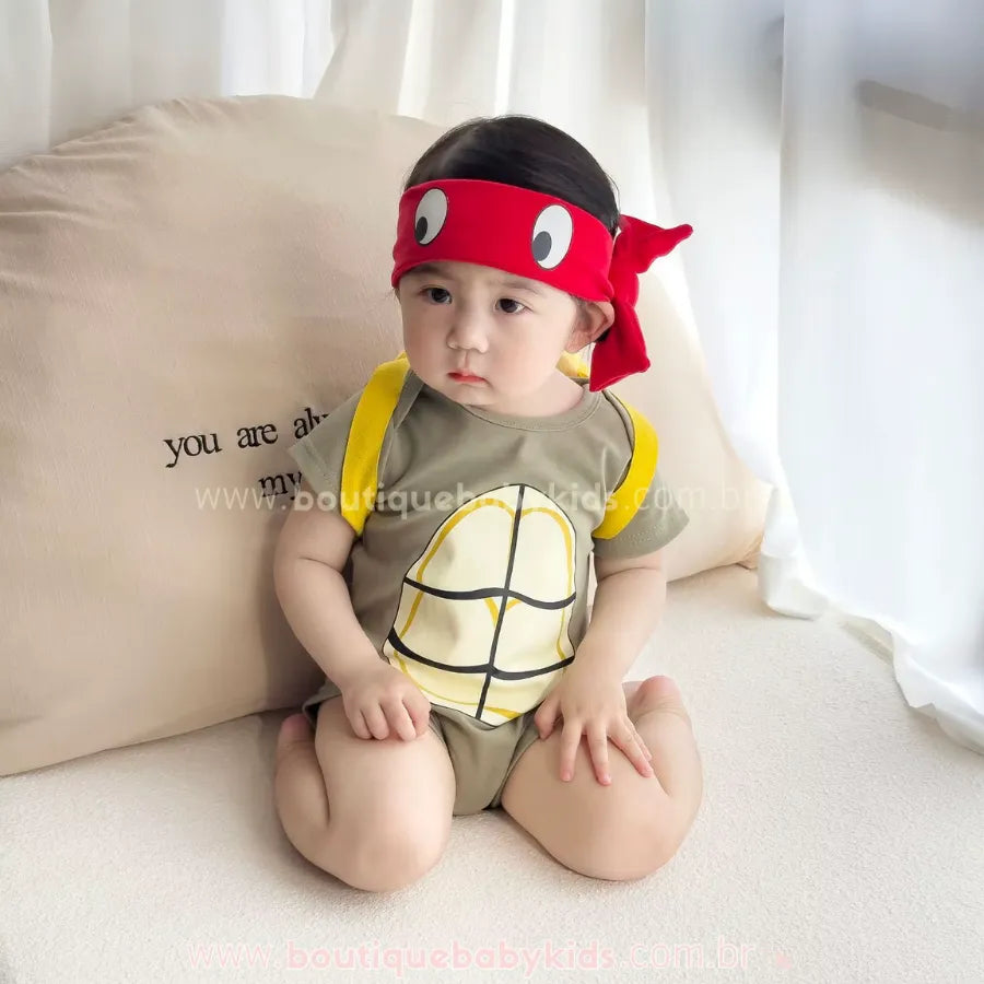 Bebê sentado vestindo Body Fantasia Tartaruga Ninja com faixa vermelha na cabeça, ideal para fotos de mesversário temático.