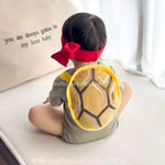 Bebê visto de costas usando o casco de pelúcia da fantasia Tartaruga Ninja, detalhando as alças macias e o acabamento confortável.