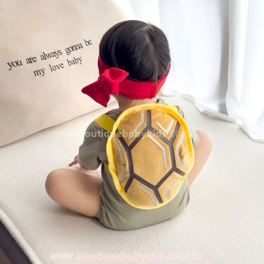 Bebê visto de costas usando o casco de pelúcia da fantasia Tartaruga Ninja, detalhando as alças macias e o acabamento confortável.
