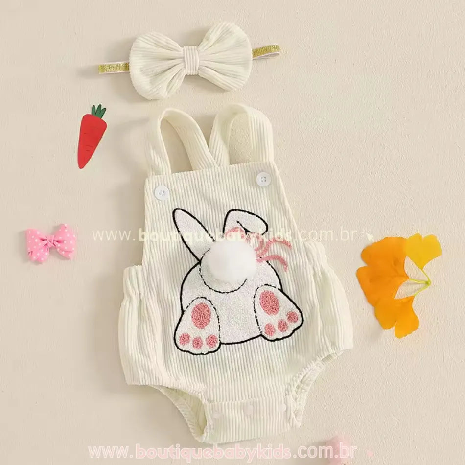 Body bebê jardineira na cor creme, disposto sobre uma superfície decorada com elementos temáticos da Páscoa, incluindo cenourinha decorativa e laço rosa. 