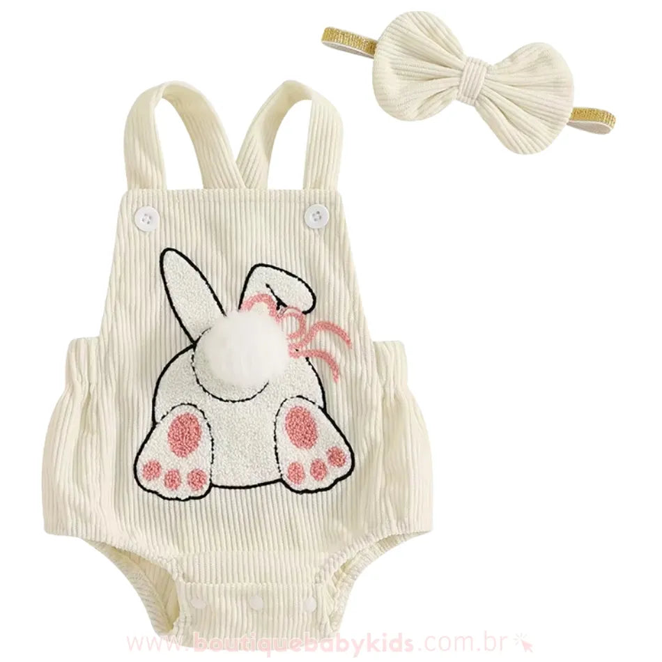 Body bebê jardineira na cor creme com bordado de coelhinho, visto de frente, acompanhado de uma faixa de cabelo combinando, com laço estruturado e detalhe dourado. 