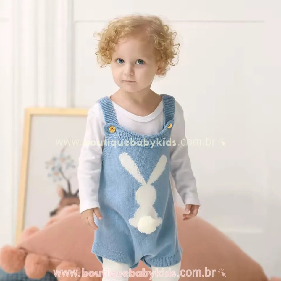 Bebê vestindo o Body Bebê Jardineira Tricot Coelhinho azul, com uma blusa branca de manga longa por baixo. O coelho branco na parte frontal se destaca contra o tricot azul, com um detalhe fofo do rabinho em pompom. O bebê está de pé em um ambiente claro e aconchegante. 