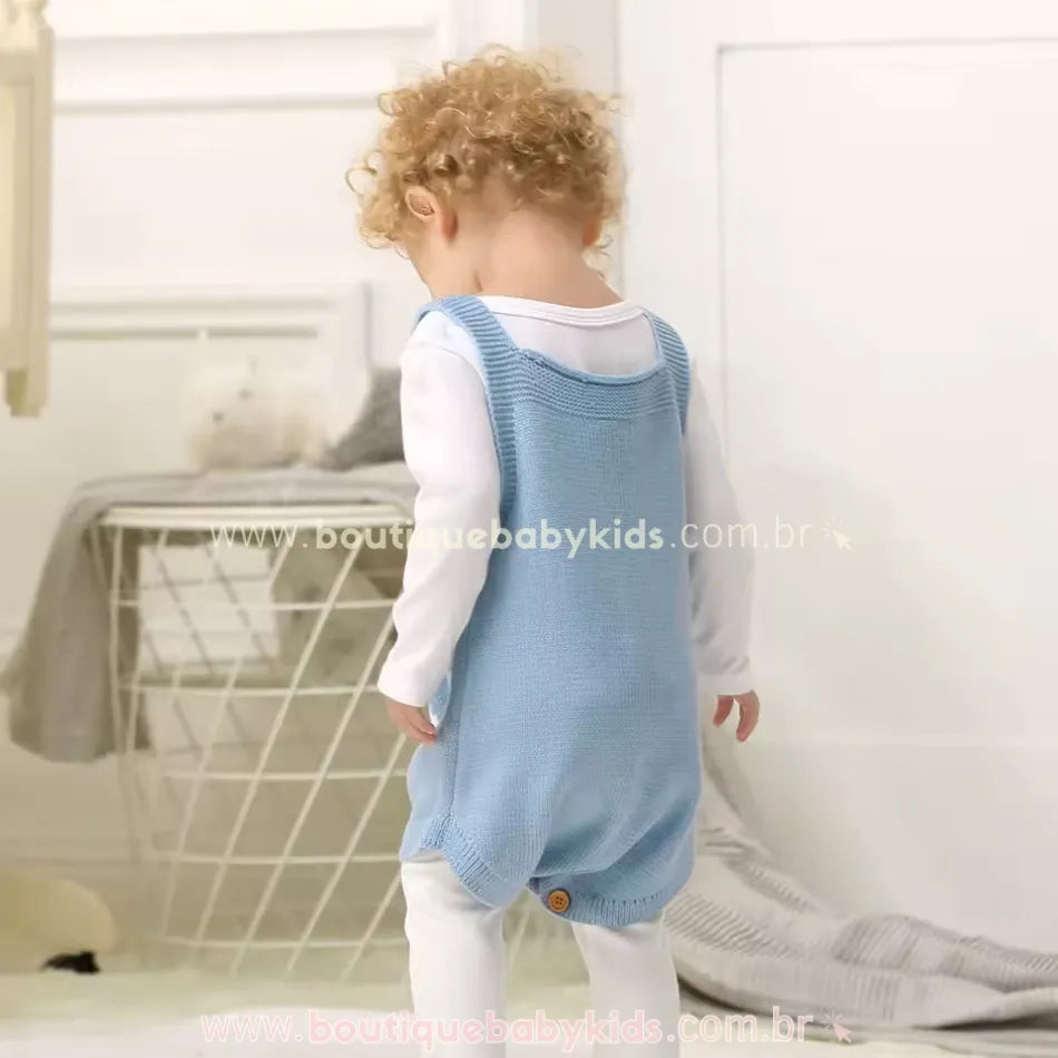 Bebê de costas, usando o Body Bebê Jardineira Tricot Coelhinho azul. A peça tem um acabamento impecável, com um botão central na parte inferior das costas para um detalhe extra. O bebê está em um ambiente aconchegante, vestindo uma blusa branca de manga longa por baixo do body. 