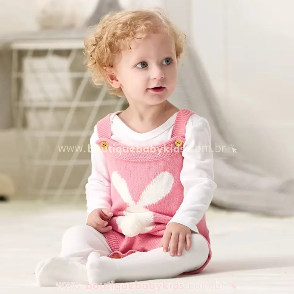 Bebê de cabelos cacheados sentado no chão, vestindo o Body Bebê Jardineira Tricot Coelhinho rosa. A peça em tricot tem uma estampa de coelho na parte frontal e um rabinho de pompom branco. O bebê usa blusa e meia-calça branca, criando um visual delicado e aconchegante. 