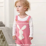 Bebê de cabelos cacheados vestindo o Body Bebê Jardineira Tricot Coelhinho na cor rosa, com uma blusa branca por baixo. O body de tricot possui alças ajustáveis com botões e um coelhinho branco tricotado na parte frontal com um pompom macio representando o rabinho. O bebê está de pé, olhando para cima com um olhar curioso. 