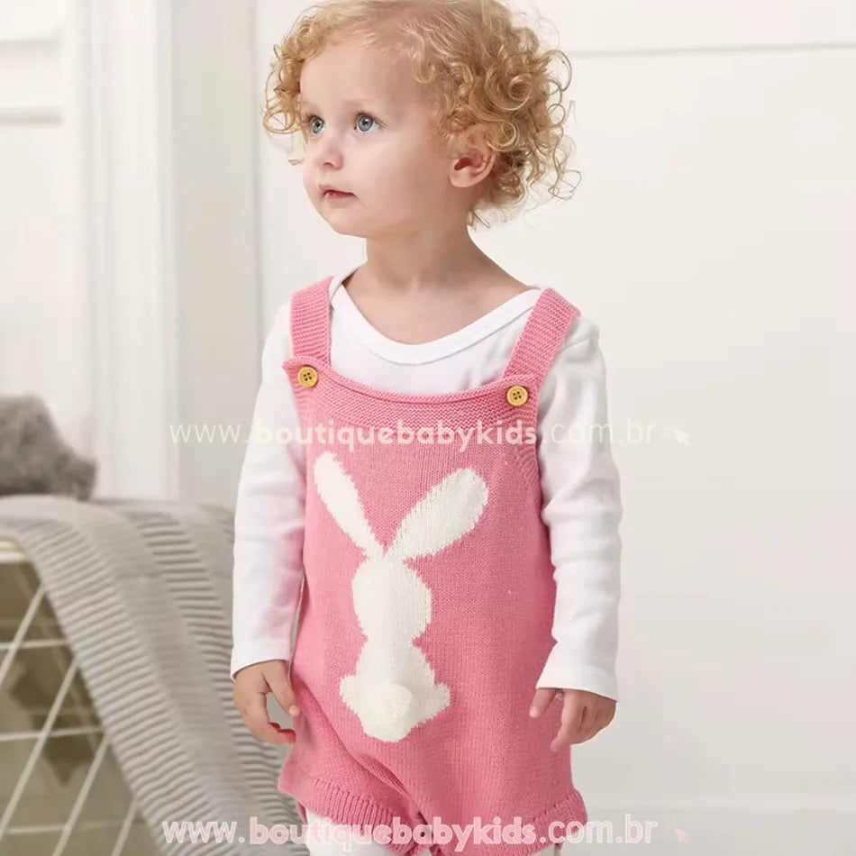 Bebê de cabelos cacheados vestindo o Body Bebê Jardineira Tricot Coelhinho na cor rosa, com uma blusa branca por baixo. O body de tricot possui alças ajustáveis com botões e um coelhinho branco tricotado na parte frontal com um pompom macio representando o rabinho. O bebê está de pé, olhando para cima com um olhar curioso. 