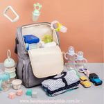 Mochila maternidade Insular Travel aberta e disposta sobre uma mesa, rodeada por mamadeiras, brinquedos e roupas, destacando seu amplo espaço para todos os itens essenciais do bebê.