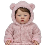 Foto de estúdio com fundo branco mostrando o rosto da Boneca Bebê Reborn Amira de frente. Ela está com o capuz de ursinho rosa levantado, destacando seus olhos claros e expressão angelical.
