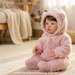 Boneca Bebê Reborn Amira de 50 cm sentada em um tapete felpudo bege. Ela veste um macacão rosa de pelúcia estilo ursinho com capuz e olha ligeiramente para o lado em um ambiente iluminado.
