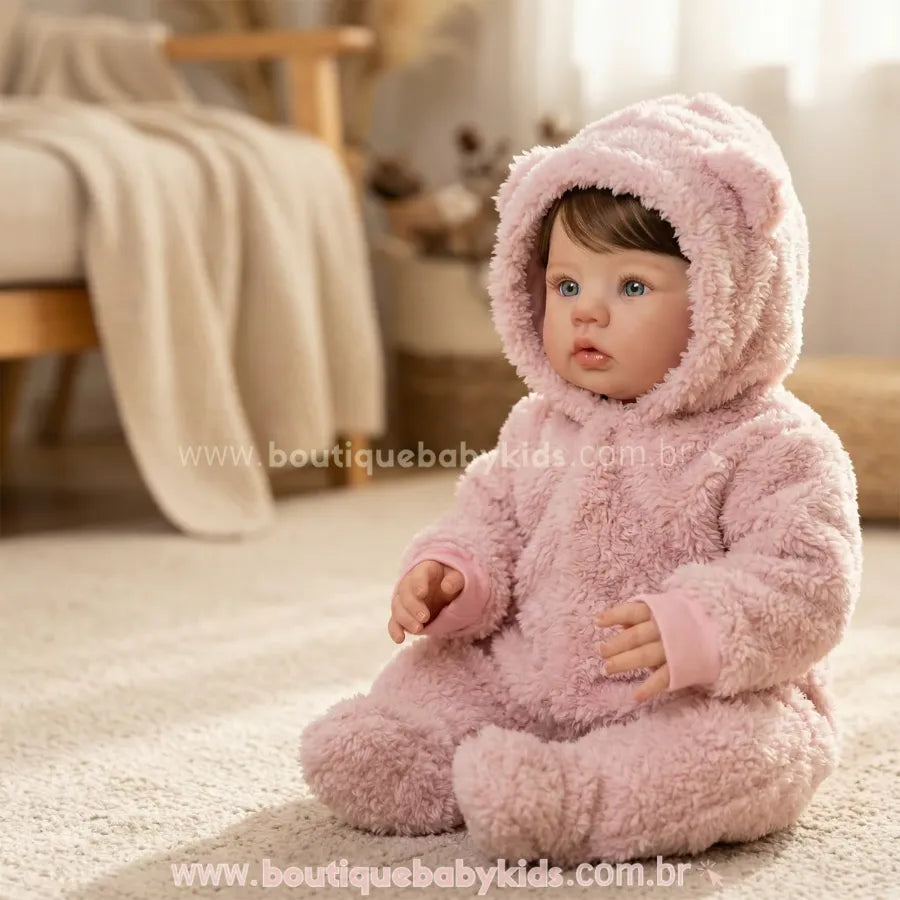 Boneca Bebê Reborn Amira de 50 cm sentada em um tapete felpudo bege. Ela veste um macacão rosa de pelúcia estilo ursinho com capuz e olha ligeiramente para o lado em um ambiente iluminado.