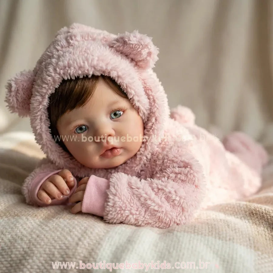 Bebê Reborn Amira deitada de bruços sobre uma manta xadrez. A boneca usa roupa de ursinho rosa com capuz e olha para a câmera, destacando seus olhos azuis realistas e cabelo castanho.