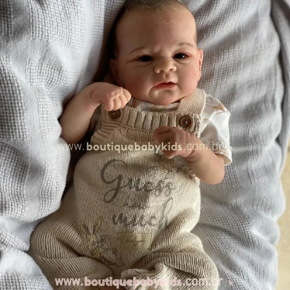 Boneca Bebê Reborn Realista Menino Bryan de corpo inteiro deitada sobre manta cinza.