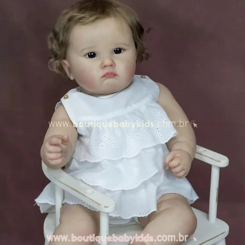 Boneca bebê reborn Catharina sentada em cadeira branca, com vestido branco de babados e expressão serena.