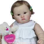 Boneca bebê reborn Catharina com fundo branco, vestindo vestido branco e segurando ursinho de pelúcia com coração rosa "Special Mum".