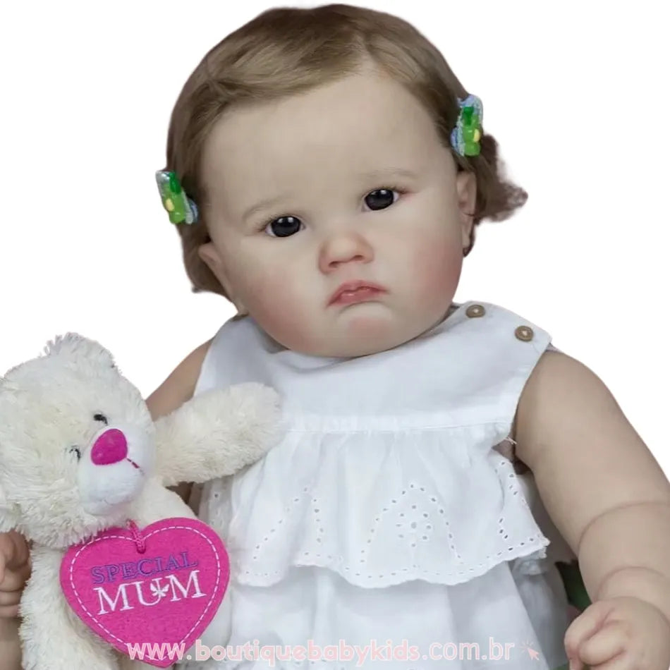 Boneca bebê reborn Catharina com fundo branco, vestindo vestido branco e segurando ursinho de pelúcia com coração rosa "Special Mum".