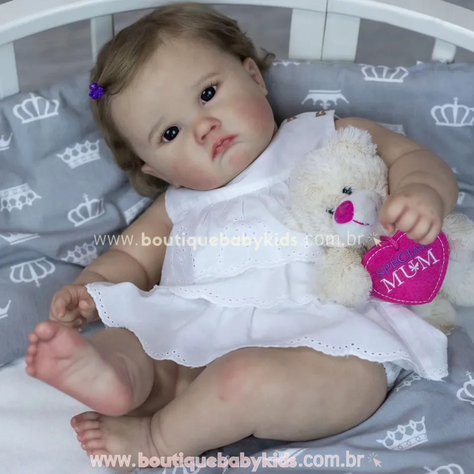 Boneca bebê reborn Catharina reclinada em berço, com vestido branco e ursinho rosa nos braços, olhando para a câmera.